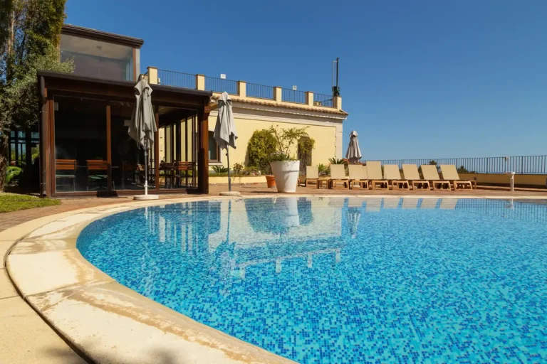 Nell'immagine, la piscina dell'Hotel Santa Caterina, con alcuni lettini sulla destra e una veranda sulla sinistra. Davanti ci sono due ombrelloni bianchi chiusi.
